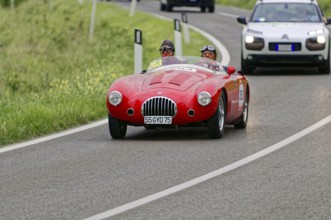 A red vintage racing car drives along green fields on a winding country road during a rally,
