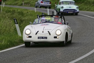 A white Porsche convertible with two occupants drives on a winding country road in front of a green