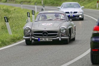 A silver vintage Mercedes car drives in front on a road, followed by another car, vintage car, car