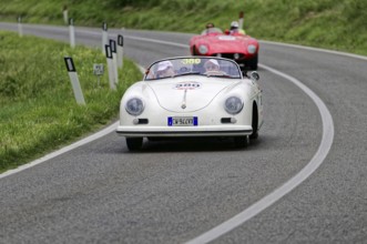 A white Porsche convertible in classic car style drives on a winding country road in front of a red