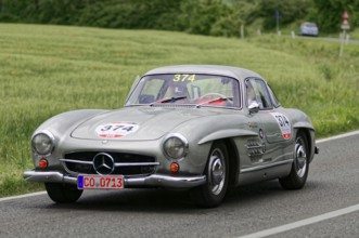 A silver Mercedes vintage car with racing stickers drives on a road in natural surroundings,