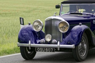 An elegant purple vintage car on a road, highlighted front headlights, surrounded by green fields,