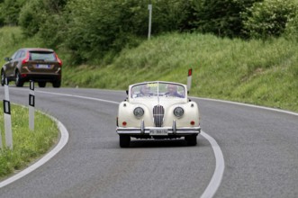A classic convertible drives along a winding country road surrounded by green countryside, classic