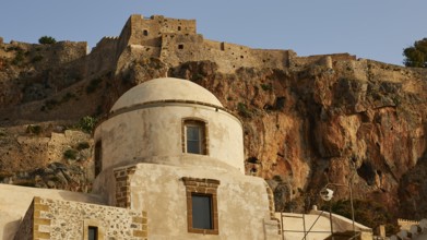 Soft morning light, historic buildings and fortress elements in front of an impressive rock wall,