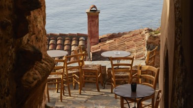 Soft morning light, narrow alley with café terrace and view of the blue sea through tiled roofs,