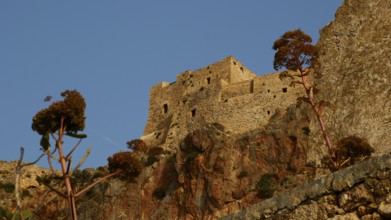 Soft morning light, old stone fortress on rocks with plants, under clear sky, Monemvasia,