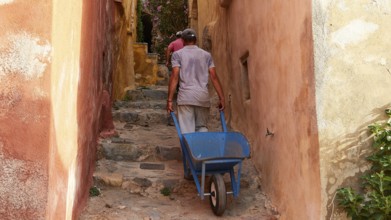 A man pulls a wheelbarrow up a narrow, colorful alley, Monemvassia, Malvasy, small Byzantine town,