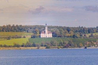 A church on a hill surrounded by vineyards and forest, with a view of a lake under a colorful