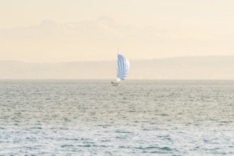 Small sailboat with blue and white sail on a quiet lake against a mountain backdrop, boat trip,