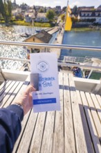 Hand holding a brochure in front of a pier with a view of water, boat trip, Lake Constance, Germany