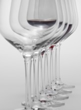 Some wine glasses standing consecutive in a row in light grey back, one partly filled with red wine