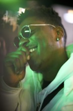 Greenlit African man wearing sunglasses at night as shade of anonymity, laughing, happy, giggle,