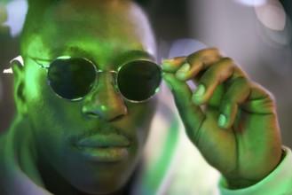 Greenlit African man wearing sunglasses at night as shade of anonymity, coolness, approved