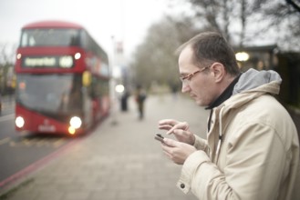 Man typing on smartphone next to double-deck bus in London, red bus, checking timetable on mobile