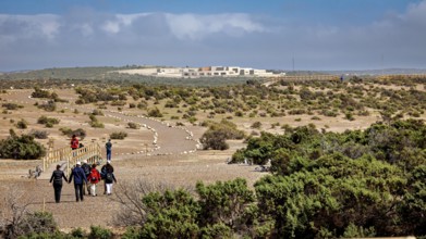 Group of people hiking on a trail through a vast desert landscape towards a complex of buildings,