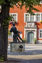 A statue in a square in front of a red, historic building with green door and blue windows, Johann