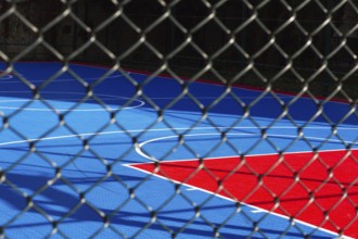 Brightly colored basketball court features blue and red sections, seen through a chain-link fence.