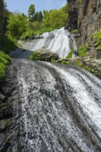 An extensive waterfall flows over rocks nestled in a green, sunny landscape, people at Jermuk