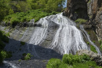 A clear waterfall flows over rocks surrounded by lush green plants in sunny nature, Jermuk