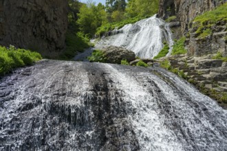 A powerful waterfall pours over steep rocks surrounded by green vegetation in the sun, Jermuk