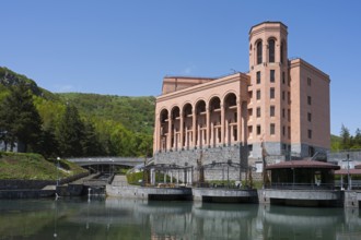 Large pink building with pillars on a river with a bridge surrounded by green hills and clear sky,