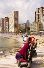 Two people in motorized wheelchairs, promenade on the beach, view of the high-rise skyline, Little