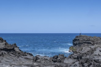 An angler stands on a massive rock on the black lava coast and looks out at the roaring Indian