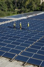 Engineers wearing safety gear are walking along rows of extensive solar panels on a rooftop,