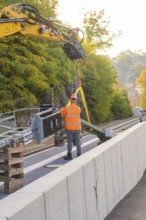 Excavator lifts objects on rails, construction worker wearing orange outfit, city in the