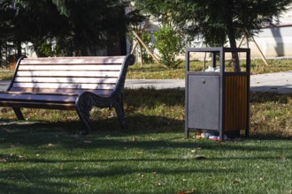 A wooden bench sits alongside a waste bin in a well-kept park. Lush green grass surrounds the