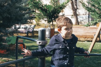 A young boy is sitting on a playground structure in a park, surrounded by trees and grass. He looks