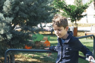 A young boy sits on a playground railing in a sunlit park, surrounded by greenery and trees. He
