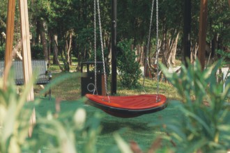 A vibrant red swing hangs suspended in a green park, surrounded by tall trees and wooden benches.