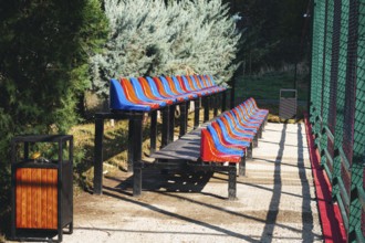Brightly colored benches line a path beside a sports field in a park. Lush greenery surrounds the