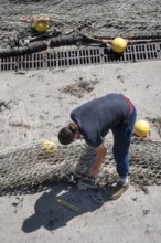 Seen from above, a fisherwoman in blue pants bends over a large fishing net spread out on the dry
