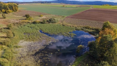 A quiet pond surrounded by meadows and trees. The colors of autumn are reflected while the sky is