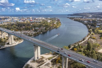 Aerial view of a bridge over a river with cars and a boat, surrounded by urban landscape. Aerial