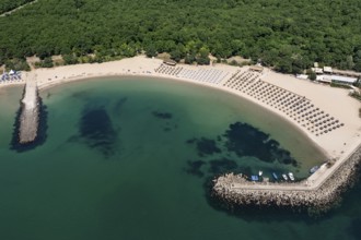 Beach lined with umbrellas and adjacent to lush greenery beside calm waters. Aerial view to