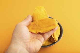 Dried Mango in blue ceramic bowl with hand on orange pastel paper background. Side view, close up.