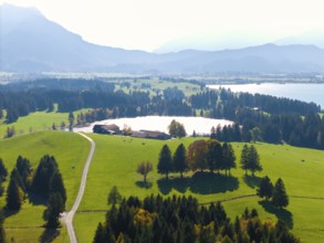 Wide, green landscape with wooded areas, lake and mountain view under blue sky, farm, near Füssen,