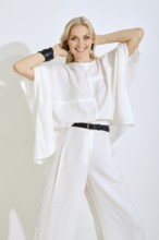 A woman stands with her arms raised, showcasing her fashionable white ensemble. She has a bright