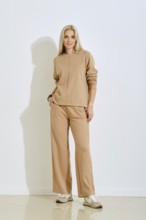 A woman stands confidently in a casual beige outfit featuring a sweater and loose pants. She poses