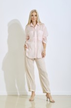 A blonde woman stands in a light pink shirt and beige pants. Her long hair flows down, and she