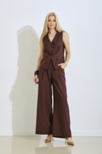 A woman stands elegantly in a chic brown tailored outfit that includes a fitted vest and wide-leg