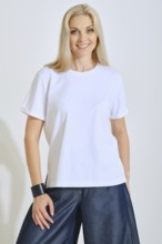 A woman stands confidently, wearing a plain white t-shirt paired with loose denim pants. She smiles