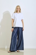 A smiling woman stands confidently in a bright studio space, wearing a simple white t-shirt and