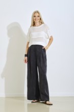 A model stands confidently indoors wearing a light top and loose black pants. The setting features