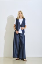 A woman stands confidently in a modern studio dressed in a denim vest and wide-legged pants. The