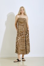 A woman stands confidently in a brown sundress featuring a unique white pattern. The setting is a