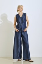A woman stands gracefully in a stylish blue denim vest and wide-legged pants. She is indoors,
